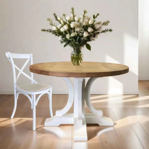 An antique round oak kitchen table with a white pedestal base holds a glass vase of flowers. A woven-seat chair sits next to it on a wooden floor.