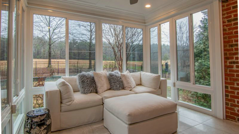 A sunroom features a white Hoolnn sectional with grey pillows and ottoman, adding texture. Trees and a fenced yard create a serene view.