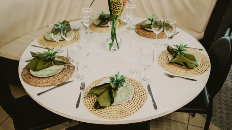 A round dining table set for six features Hoolnn's decor: woven placemats, green napkins, wine glasses, and a plant centerpiece.