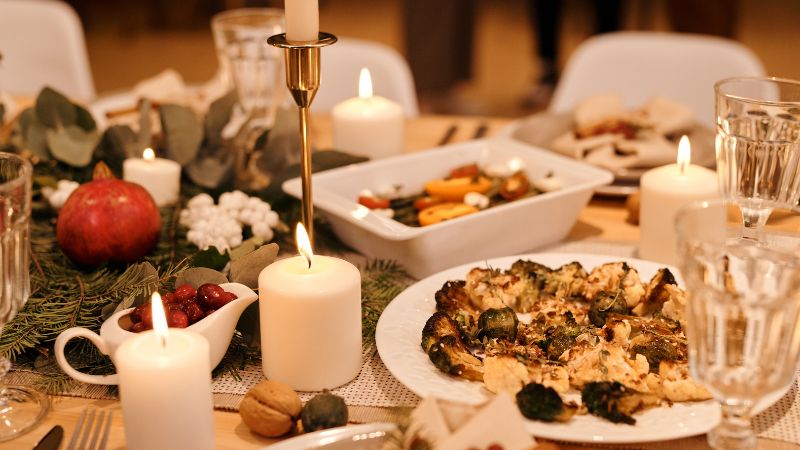 Hoolnn's round table decor showcases greenery and candles, with roasted veggies, a pomegranate, and elegant glassware for a festive dinner.