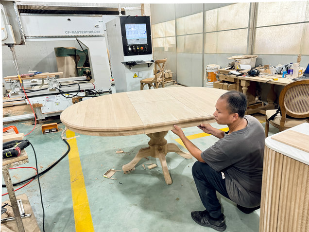 Someone examines an oval wooden table in a workshop filled with tools and machinery.