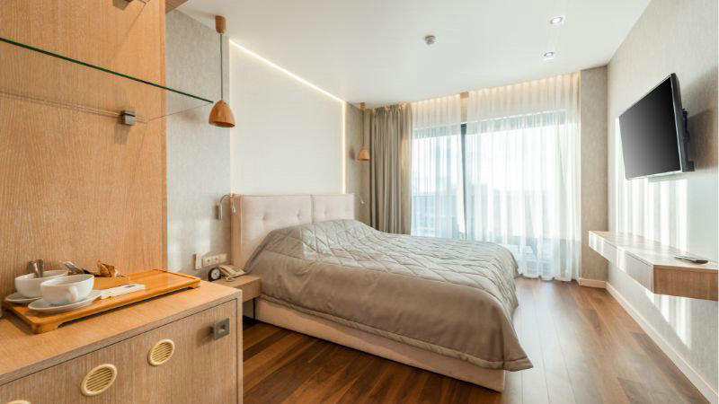 The modern hotel room has a HOOLNN platform bed, a wall-mounted TV, sleek wooden flooring, and bright light through sheer curtains.