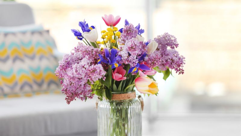 A Hoolnn glass vase of lilacs, tulips, and irises graces a round table, enhancing the decor in a bright room with charm.