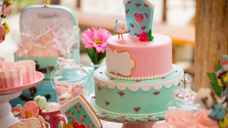 A vibrant two-tier cake with floral decor and a bird topper, surrounded by candies and pink cupcakes, elevates your life.