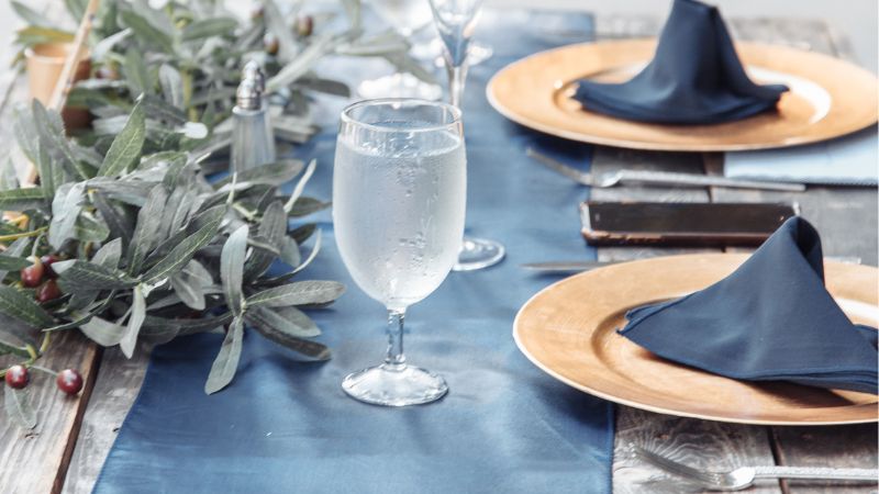 Enhance your dining with Hoolnn's round table decor: gold-rimmed plates, navy napkins, a glass of water, and lush greenery on a blue runner.