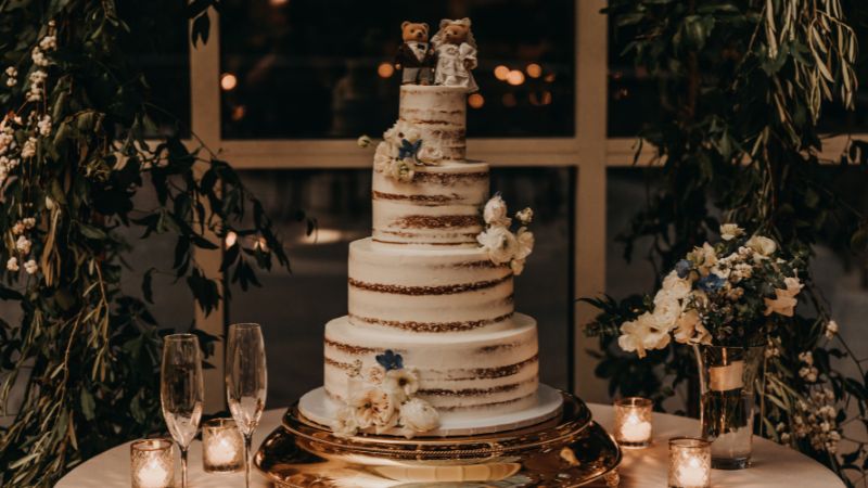 A three-tier naked wedding cake with flowers and bear toppers graces an elegant table, surrounded by greenery, candles, and champagne flutes.