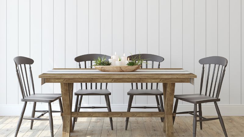 A wooden table with four dining chairs, a striped runner, and a candle and succulent centerpiece set against a white paneled wall.