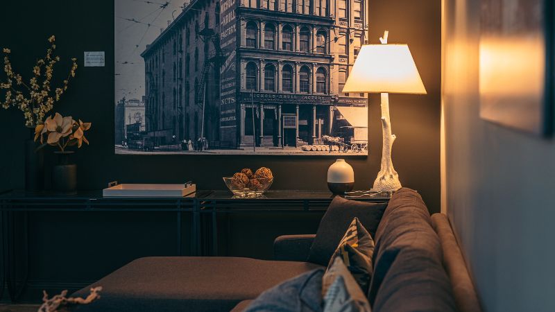 Moderna viesistaba ar brūnu sekciju, lampu uz sānu galdiņa un pilsētas ainavas fotoattēlu — ideāli piemērota ātrai lampas augstuma noteikšanai.