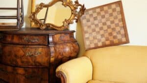 A checkered board and ornate mirror rest on an antique dresser by a yellow upholstered sofa.