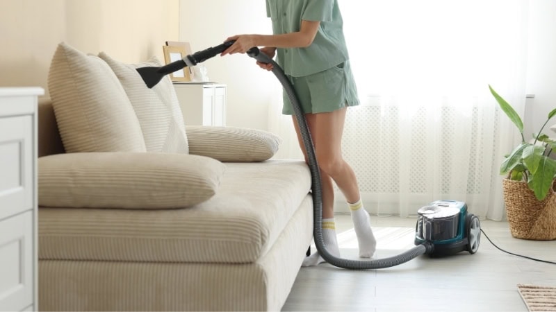 Woman vacuuming a beige linen sofa to remove dust and dirt, maintaining its cleanliness and appearance. Perfect for furniture cleaning and home care tips.