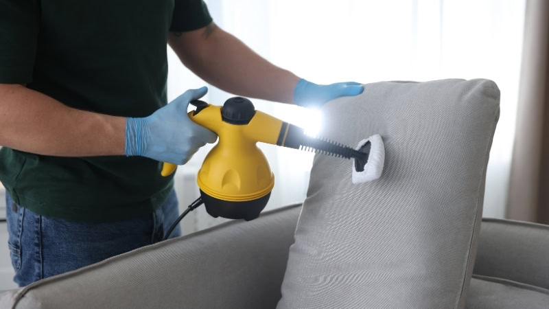 Person cleaning a light-colored linen sofa with a handheld steam cleaner, demonstrating effective upholstery cleaning methods for maintaining a fresh and beautiful linen furniture.