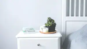 Eine gemütliche Schlafzimmerszene mit einem weißen Nachttisch, einer kleinen Topfpflanze, Kerzen und Büchern schafft eine entspannende und minimalistische Atmosphäre, die sich perfekt für einen erholsamen Schlaf eignet und als Inspiration für die Schlafzimmergestaltung dient.