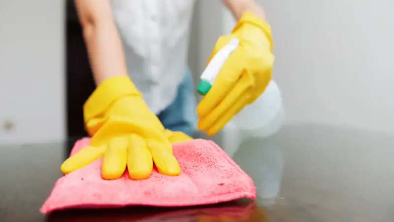 Woman cleaning antique wood furniture with a pink cloth and spray, wearing yellow gloves, demonstrating safe cleaning techniques for delicate wooden surfaces.